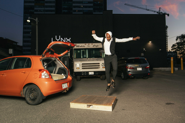 man tap dancing outside of a building beside an orange car