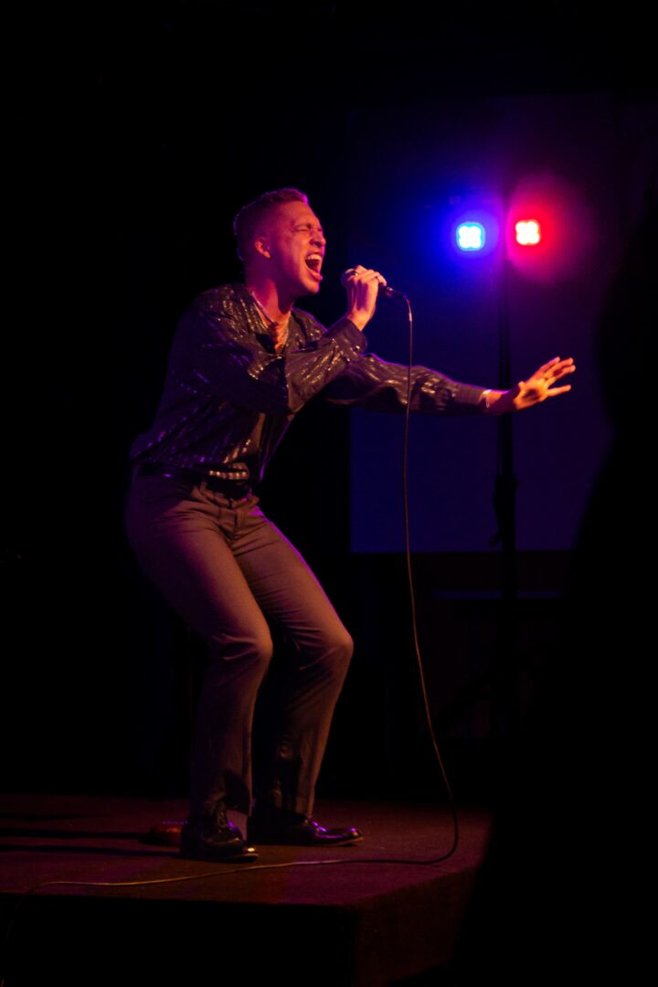 Gallery 1 - A performer (Alten Wilmot) singing into a microphone, illuminated by red and blue stage lights.